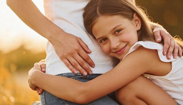 Smiling daughter hugging pregnant mother in golden meadow at sunset