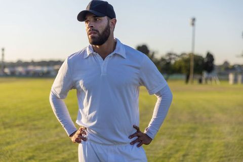 Cricket player standing on grassy field during golden hour wearing white uniform and cap