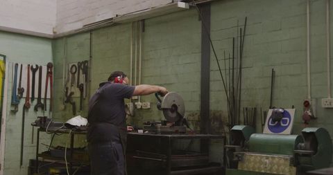 Metalworker cutting steel with chop saw in workshop wearing red earmuffs and apron