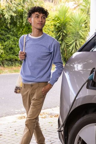Young man standing beside charging electric car with grocery bag
