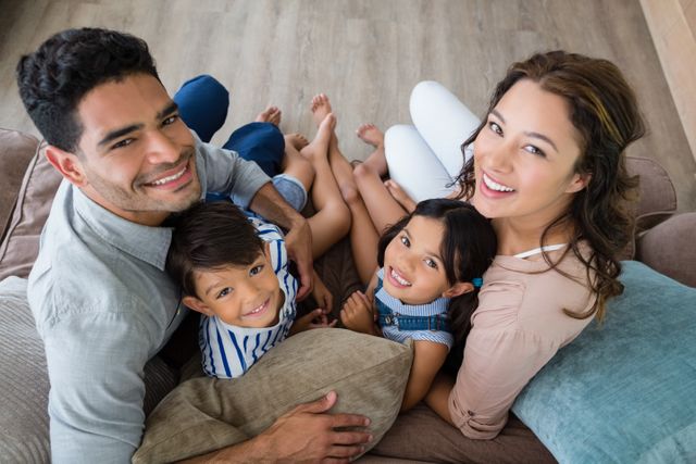Family sitting together on sofa, smiling and enjoying each other's company. Perfect for advertisements focusing on family values, home decor, and lifestyle. Ideal for parenting blogs, social media campaigns on family bonding, and brochures for family-friendly products or services.