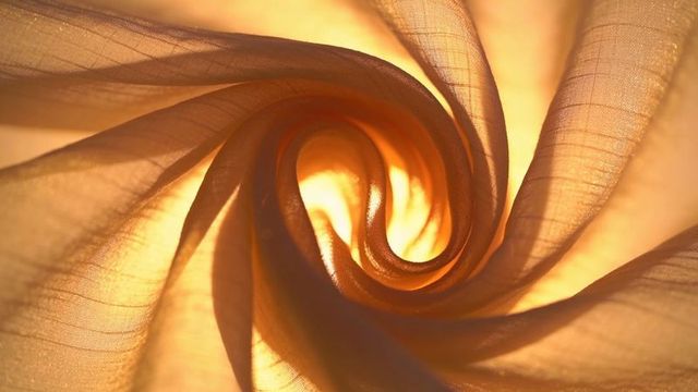 Glowing sheer fabric spiraling in golden backlight revealing woven translucent texture
