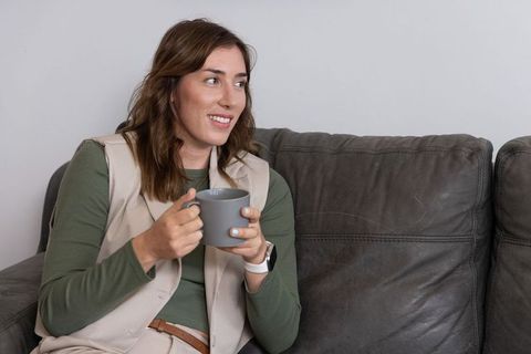 Smiling woman sipping coffee on gray leather sofa wearing vest and smartwatch
