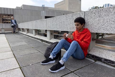 Teen sitting on urban plaza using smartphone wearing red hoodie and backpack