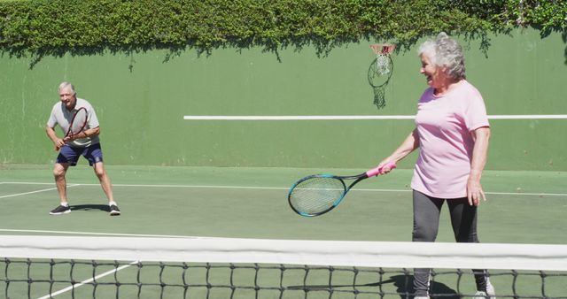 Elderly man and woman enjoying a game of tennis together on a green outdoor court. This image can be used to promote active lifestyles for seniors, fitness and health programs for the elderly, recreational sports activities, and social interaction among older adults. Great for fitness brochures, senior living publications, and sports event advertisements.