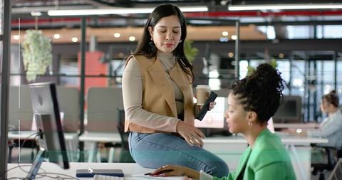 Two professional women collaborating in modern open office with laptop, coffee and monitor