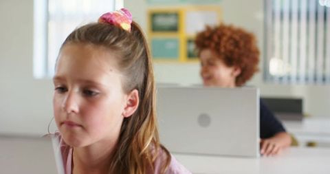 Preteen girl studying attentively while classmate using laptop in bright diverse classroom