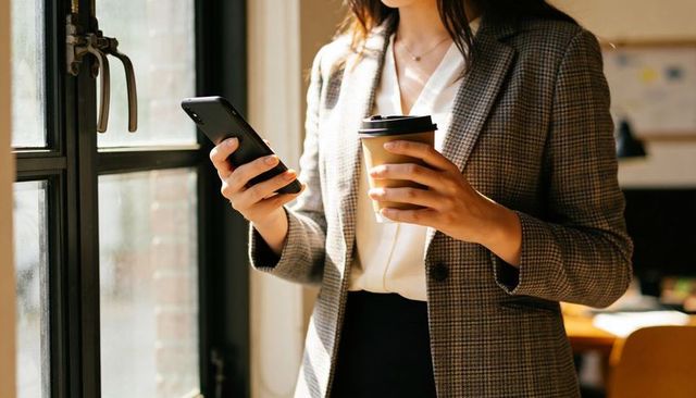 Businesswoman holding smartphone and coffee by window in modern office workspace