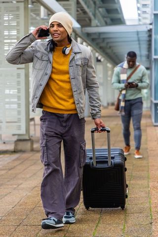 Diverse traveler pulling suitcase and talking on phone at train platform wearing beanie