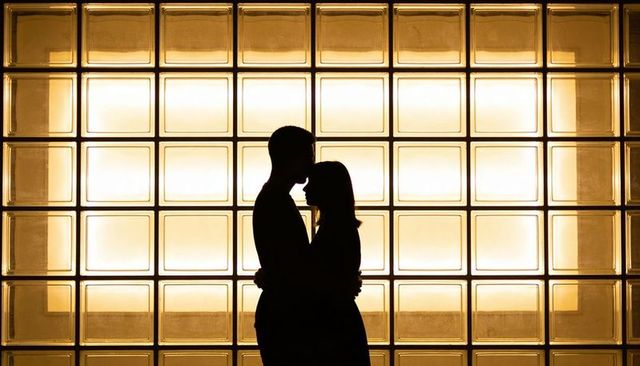 Embracing couple silhouette leaning close against warm backlit glass block grid lobby