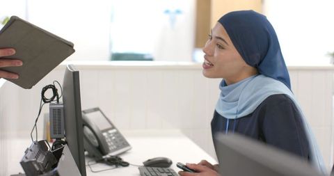 Receptionist wearing blue headscarf assisting visitor at modern medical reception desk