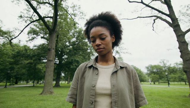 Contemplative young woman standing in urban park, gazing downward under overcast sky