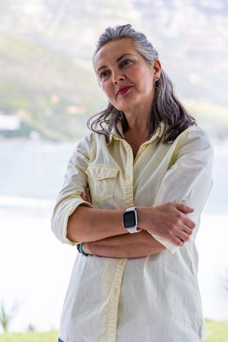 Mature woman standing arms crossed by lake wearing button-up shirt and smartwatch reflecting