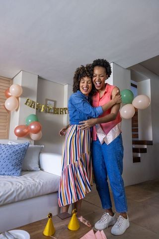 Two diverse women hugging and laughing while celebrating birthday at home with balloons