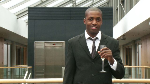 Young, stylish businessman stands confidently with glass of champagne, celebrating professional success indoors of modern office building. This uplifting and motivational scene ideal for use in articles about workplace achievements, business events, corporate culture, or promotional material for various business-related platforms or celebrations.