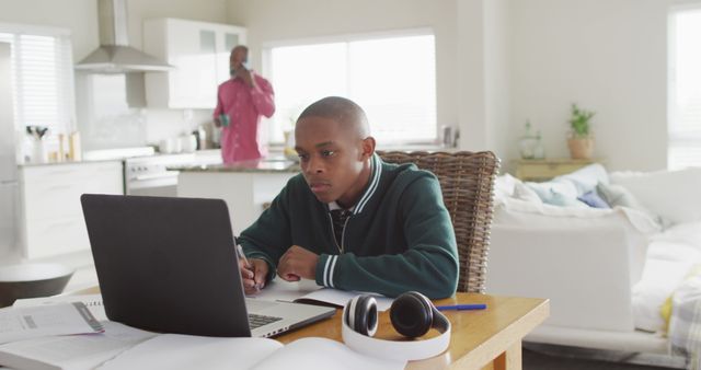 A young student is intensely focused on a laptop, seated at a desk in a home environment. Books, notebooks, and headphones are on the desk. In the background, an adult is talking on a phone in a modern kitchen, indicating a busy household. Perfect for representations of remote learning, family life, education, multitasking parents, and modern lifestyles.