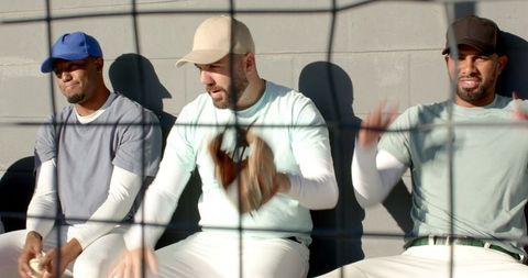 Diverse baseball teammates sitting on bench behind netting wearing caps and mitts in sunlight