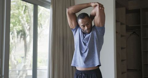 Athletic man stretching overhead by sliding glass door in sunlit minimalist bedroom
