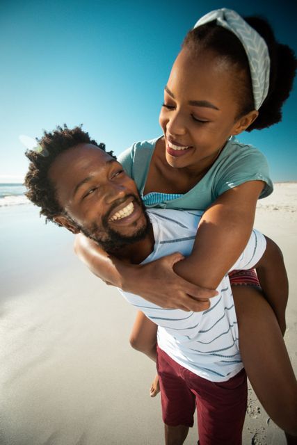 This image captures a joyful African American couple enjoying a playful piggyback ride on a sunny beach. Ideal for use in travel brochures, romantic getaway promotions, lifestyle blogs, and advertisements focusing on leisure, love, and outdoor activities.