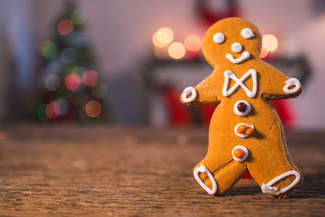 Gingerbread man decorated with icing standing on a rustic wooden table. Blurred Christmas lights and tree in the background create a festive atmosphere. Ideal for holiday greeting cards, baking blogs, festive advertisements, and Christmas-themed social media posts.