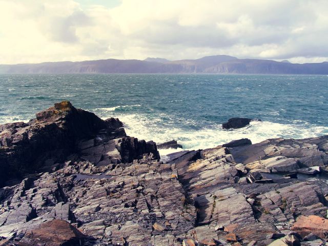 This image depicts a rugged coastline with rough waves crashing against rocks. Distant mountains are visible under a partly cloudy sky. It can be used for themes involving nature, adventure, outdoor activities, travel, and relaxation. Ideal for travel blogs, magazines, and environmental projects.