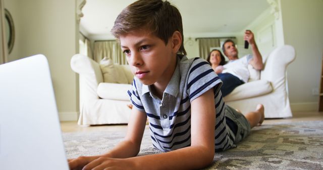 Young boy is focused on using a laptop while lying on the floor. Parents are relaxing on a couch in the background. This image can be used for family-oriented content, technology lifestyle, remote learning, and casual home environment themes.