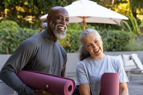 Mature diverse couple holding yoga mats and smiling on sunlit backyard patio