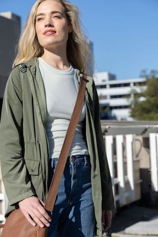 Sunlit young woman standing on rooftop wearing olive parka, ribbed top and crossbody bag
