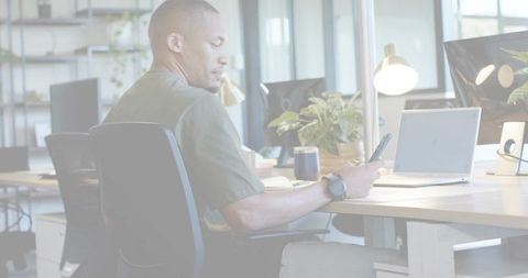 Checking smartphone at modern shared desk with laptop, plants and lamp in daylight office