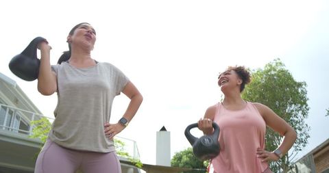 Two diverse women lifting kettlebells and laughing in backyard wearing smartwatches