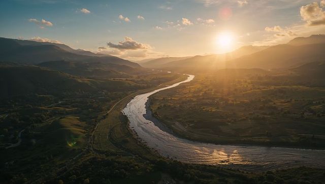 Gliding golden-hour aerial of meandering river reflecting sunlight across rural valley hills