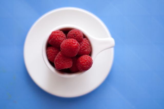 Fresh raspberries fill a white cup viewed from overhead, contrasted against a bright blue background. Great for promoting healthy eating, fruit recipes, summer snacks, minimalist designs, or a colorful food blog.