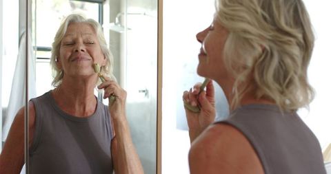 Senior woman using jade roller at mirror for daily anti-aging skincare routine