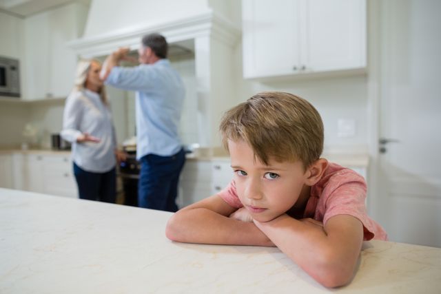 Young boy leaning on kitchen counter, looking sad while parents argue in background. Useful for illustrating family conflict, emotional distress in children, and domestic issues. Suitable for articles, blogs, and educational materials on family dynamics and child psychology.