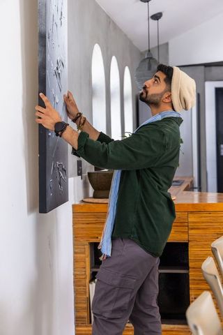 Man hanging black canvas in modern home hallway with wooden console and arched windows