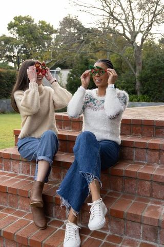 Diverse friends sitting on brick steps wearing festive holiday glasses and cozy sweaters