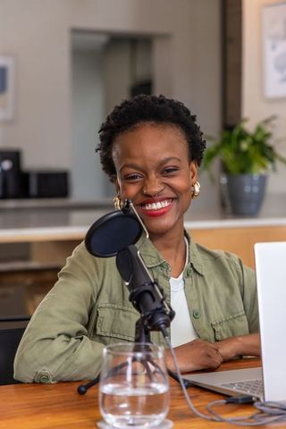 African american woman smiling while podcasting at kitchen desk with laptop and mic