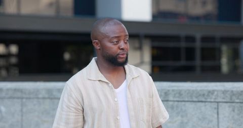 African american man standing by urban office plaza wearing beige shirt and white tee