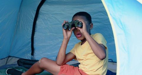 African american boy peering through binoculars inside blue tent during camping adventure