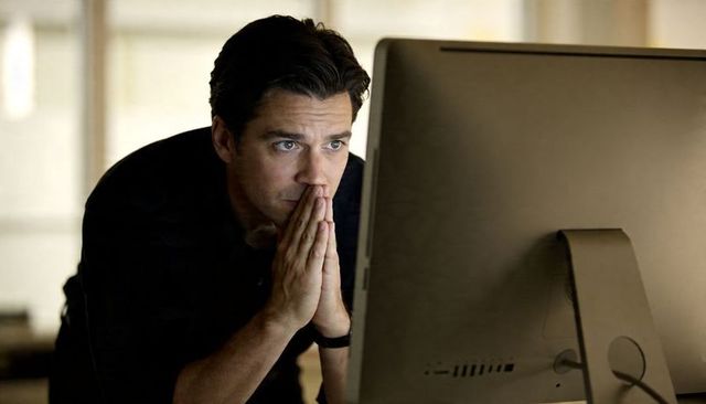 Businessman leaning over desk clasping hands intently focusing on large desktop monitor