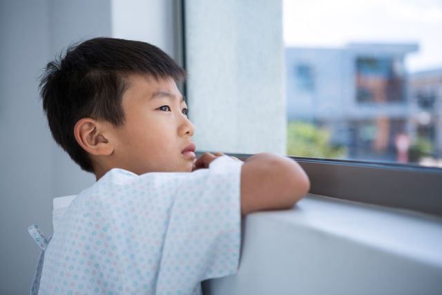 Young boy patient in hospital gown looking out window with a tensed expression. Suitable for use in healthcare, pediatric care, medical treatment, and emotional well-being contexts. Can be used in articles, blogs, and promotional materials related to children's health, hospital experiences, and patient care.