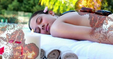 Young woman receiving hot stone massage on outdoor spa bed with candle and pink flower
