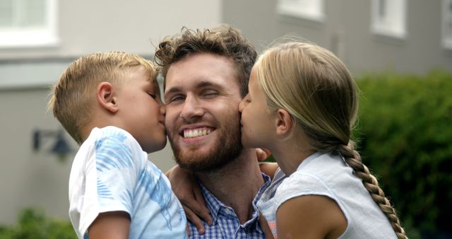 A young Caucasian man is being affectionately kissed on the cheeks by a boy and a girl, his children, with copy space. Their joyful expressions and the residential backdrop suggest a warm family moment of love and connection.