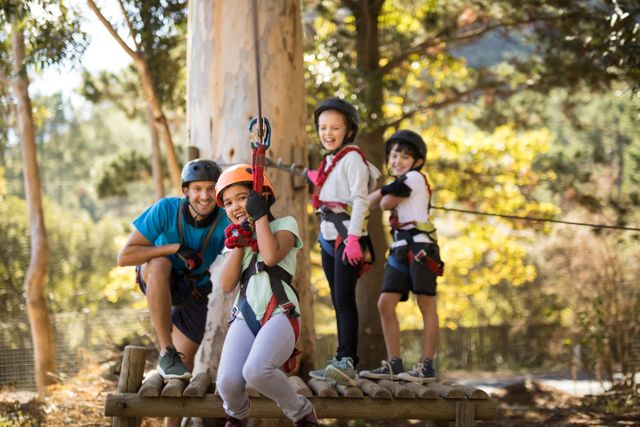 Group of children smiling and having fun while participating in a zip line adventure in the forest on a sunny day. They are wearing safety gear including helmets and harnesses. Perfect for illustrating outdoor activities, adventure sports, family bonding, summer camps, and children's recreational activities.