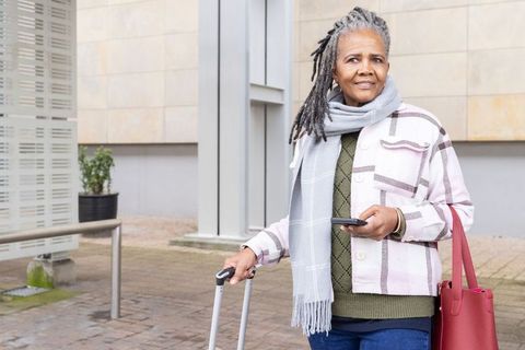 Senior african american woman traveling with rolling suitcase and smartphone in city plaza