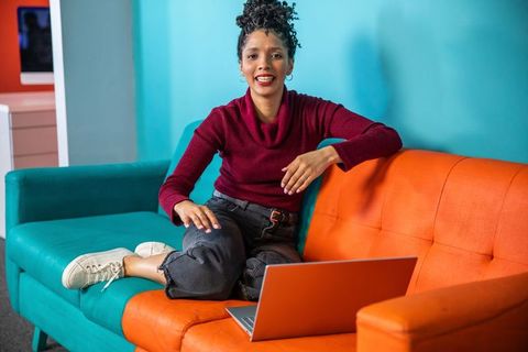 African american woman sitting on vibrant two-tone sofa working on laptop in living room