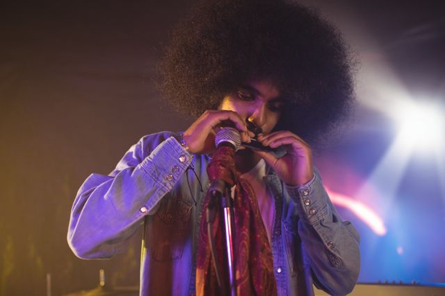 Male musician with an afro hairstyle playing a mouth organ in a nightclub. He is wearing a denim shirt and performing under colorful stage lights. This image can be used for promoting live music events, concerts, nightlife entertainment, and showcasing musical talent and creativity.