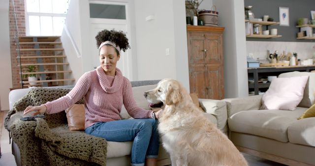 Woman sitting on couch in cozy living room, petting a golden retriever, with remote control in hand. Ideal for depicting home comfort, pet companionship, or relaxed lifestyle. Useful in advertisements for home products, pet care, or modern living spaces.