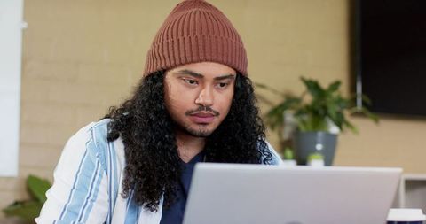 Young asian man concentrating on laptop in contemporary coworking space wearing beanie