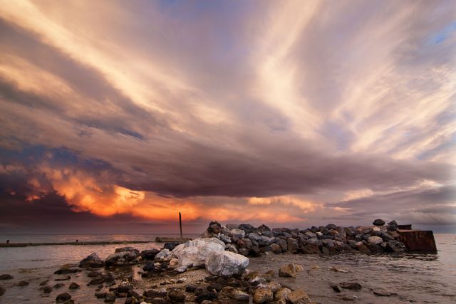 Expansive view of a dramatic sunset sky over a rocky shoreline with dark clouds illuminated by the setting sun. Ideal for use in travel brochures, nature magazines, or as part of a relaxing home or office décor theme aimed at capturing the tranquil beauty of coastal sunsets.